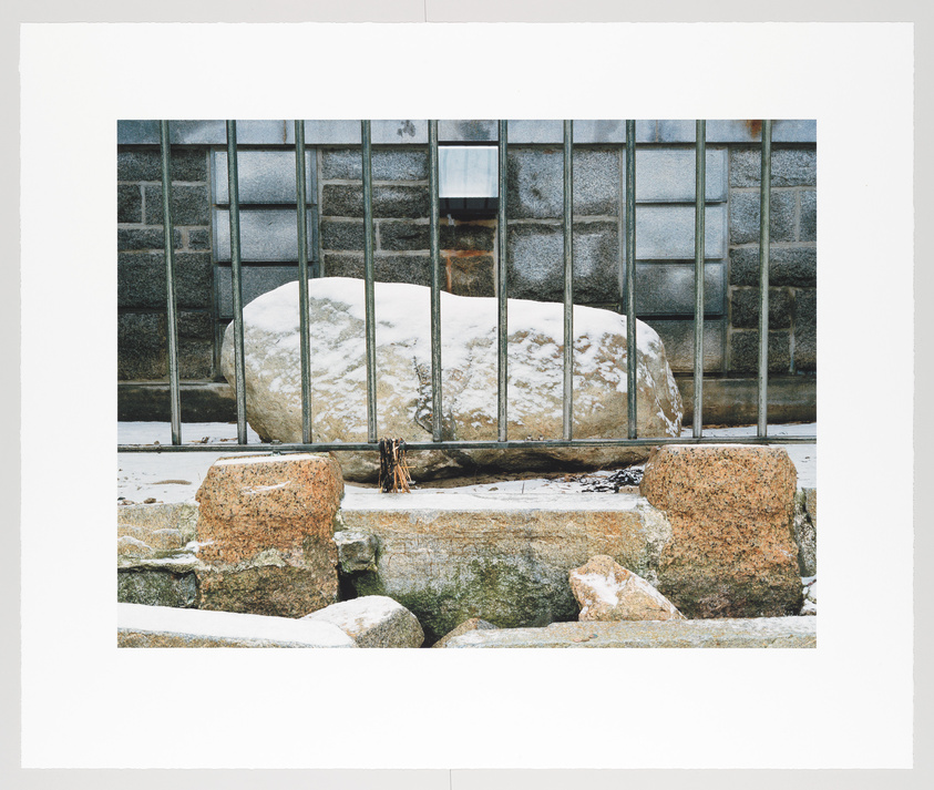 A snow-covered boulder sits behind a metal barred fence with a stone wall in the foreground, against a backdrop of a building with frosted windows.