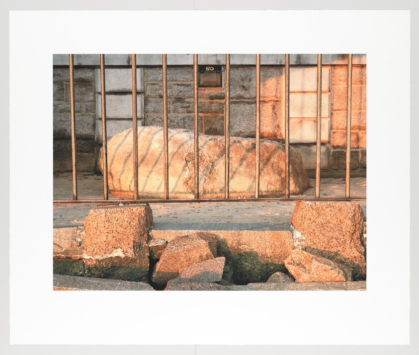 A large, weathered rock is enclosed behind a rusty metal gate with vertical bars, set against a wall with square stone tiles. In the foreground, broken pieces of concrete are scattered on the ground. The scene suggests a neglected urban space.
