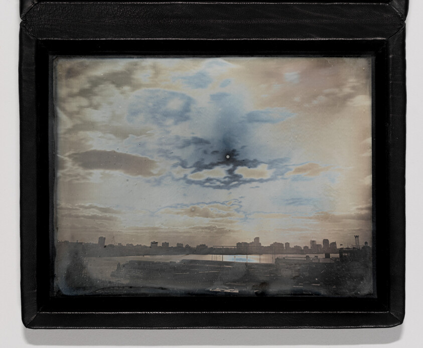 Framed vintage photograph showing a city skyline across a river under a cloudy sky with sun.