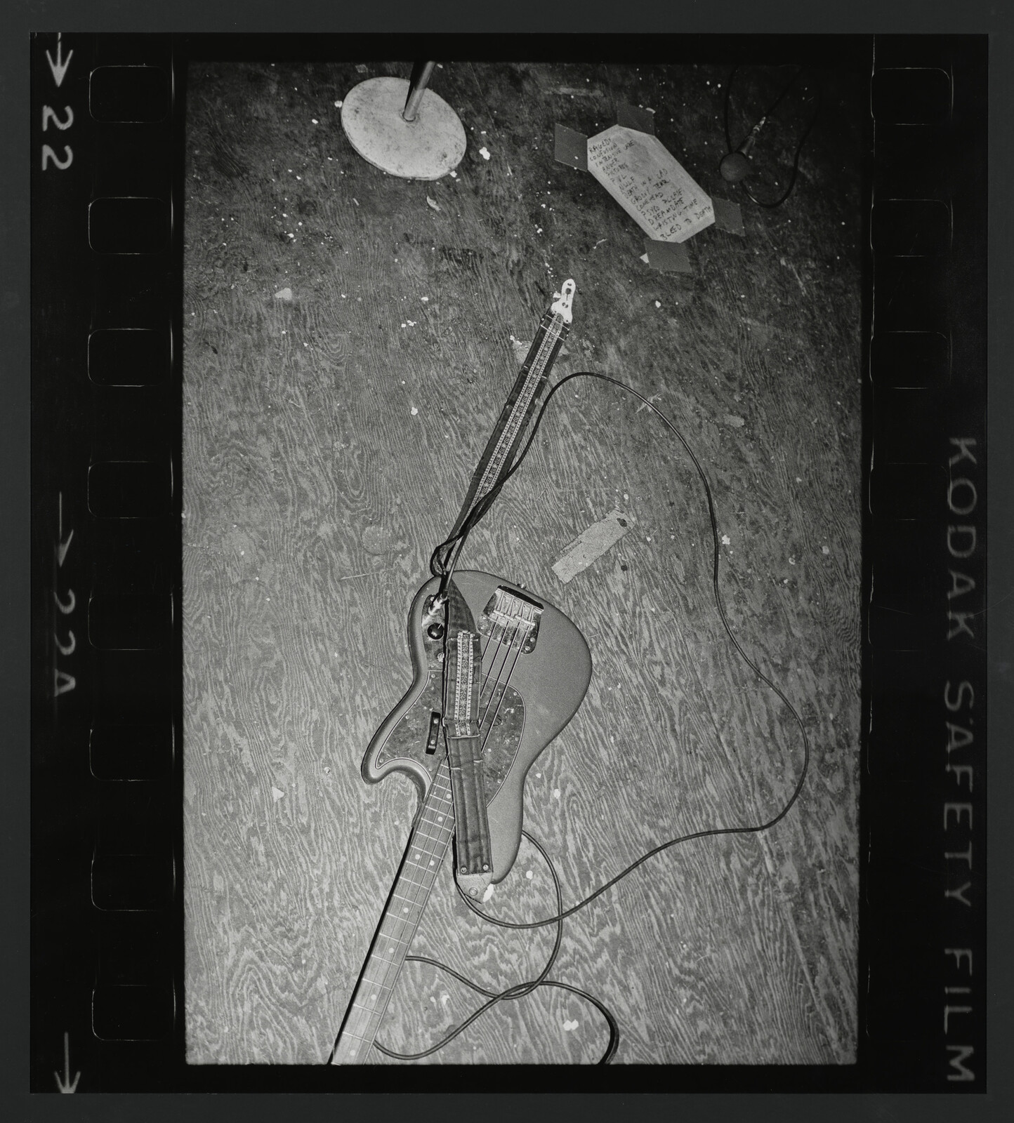 Electric bass lies on a scuffed wooden floor with cables and a taped setlist.
