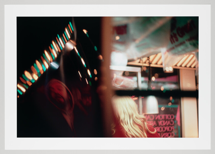 A blonde person stands near a brightly lit carnival booth reflected in a glass window.