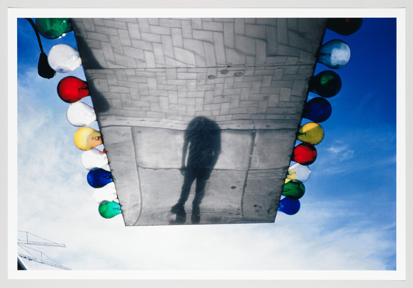 Shadow of a person reflected on a metallic canopy bordered by colorful round lights against blue sky.