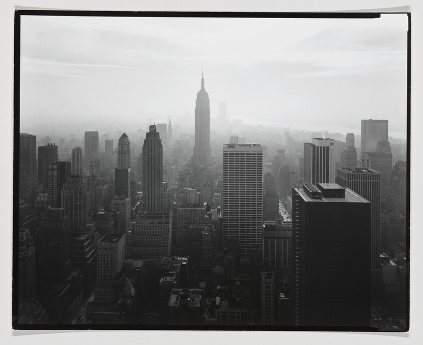 Black and white photograph of a city skyline with numerous skyscrapers, including a prominent, iconic tower in the center, shrouded in a hazy atmosphere.