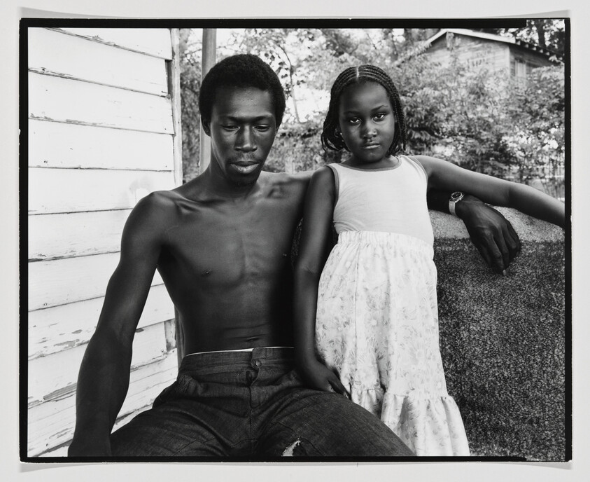 A black and white photograph featuring a shirtless man and a young girl sitting next to each other. The man, looking down, has his arm extended along the back of a bench, while the girl, wearing a white floral dress, stands with her arm resting on his, looking directly at the camera with a serious expression. They are outdoors, with trees and a white wooden structure in the background.