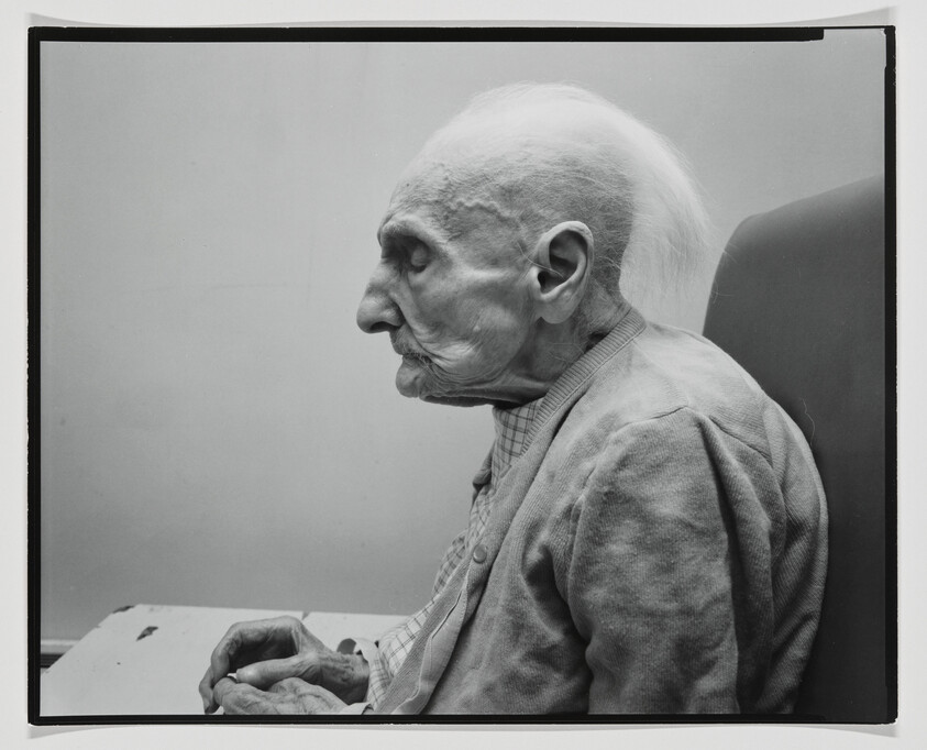 Very old woman sitting in profile with hands folded, eyes closed, wearing a cardigan.