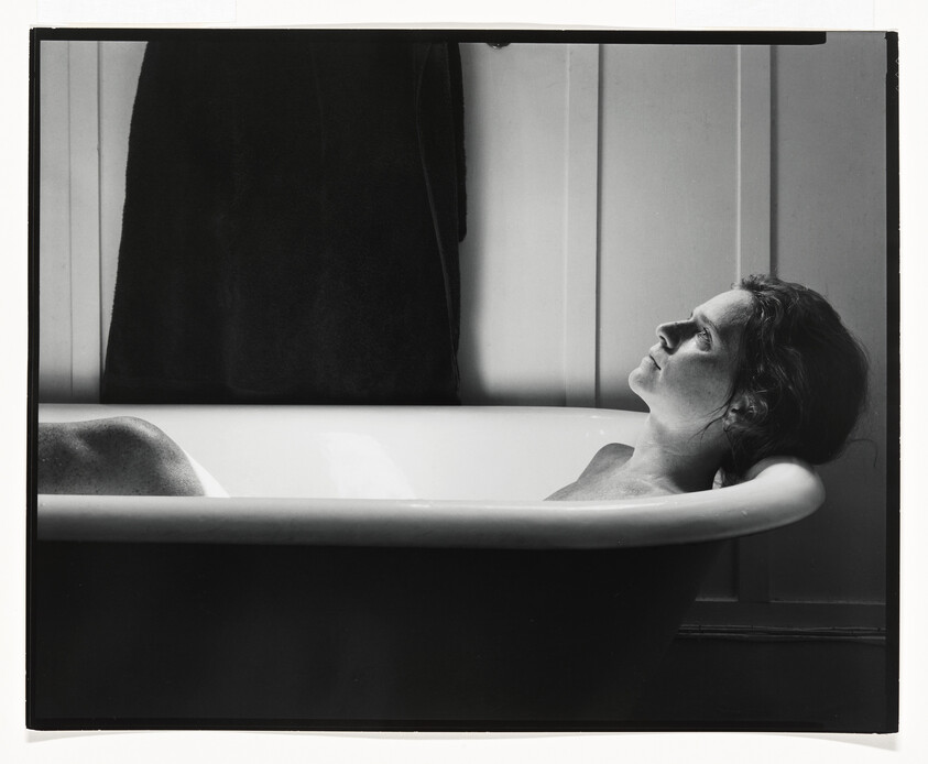 A woman reclines in a bathtub, resting her head and looking upward thoughtfully.