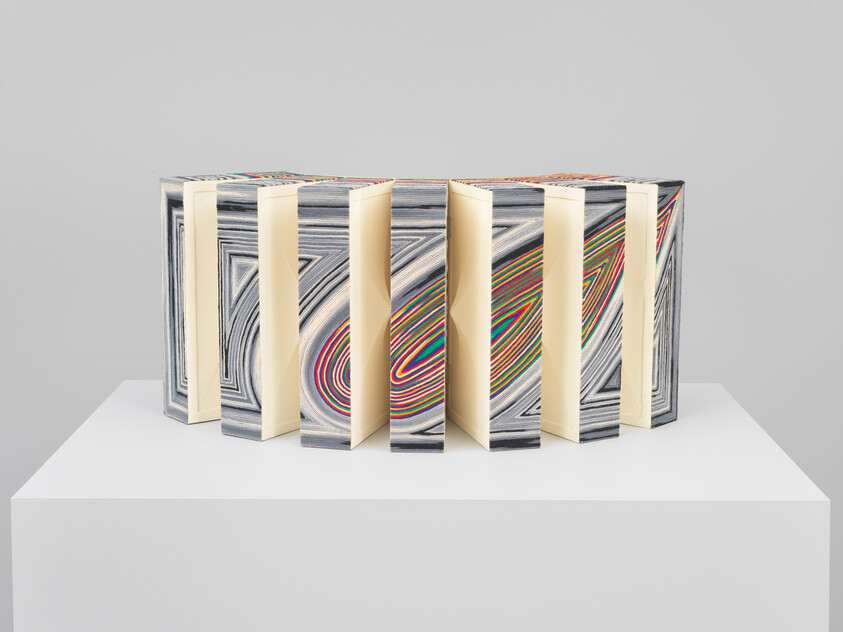 A folded accordion-like sculpture made of layered colorful paper stands on a white pedestal.