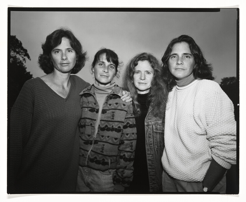 Four women standing closely together, posing for a black and white photograph. They are dressed in casual attire, with two wearing sweaters, one in a patterned fleece, and one in a denim jacket. They appear to be outdoors, with trees faintly visible in the background. The image captures a sense of camaraderie or family among the group.