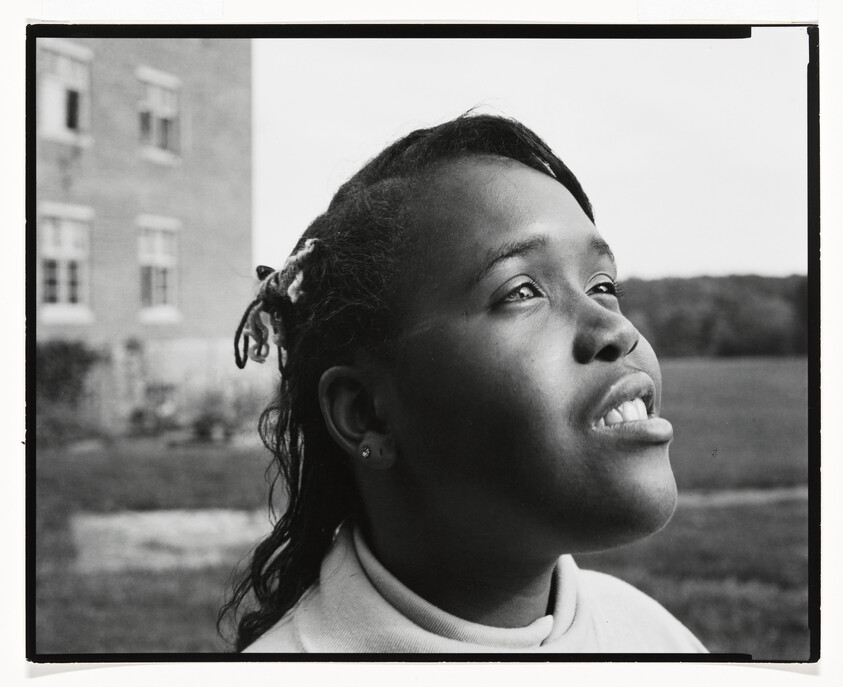 Young woman looking upward with hopeful expression outdoors near a building and open field.