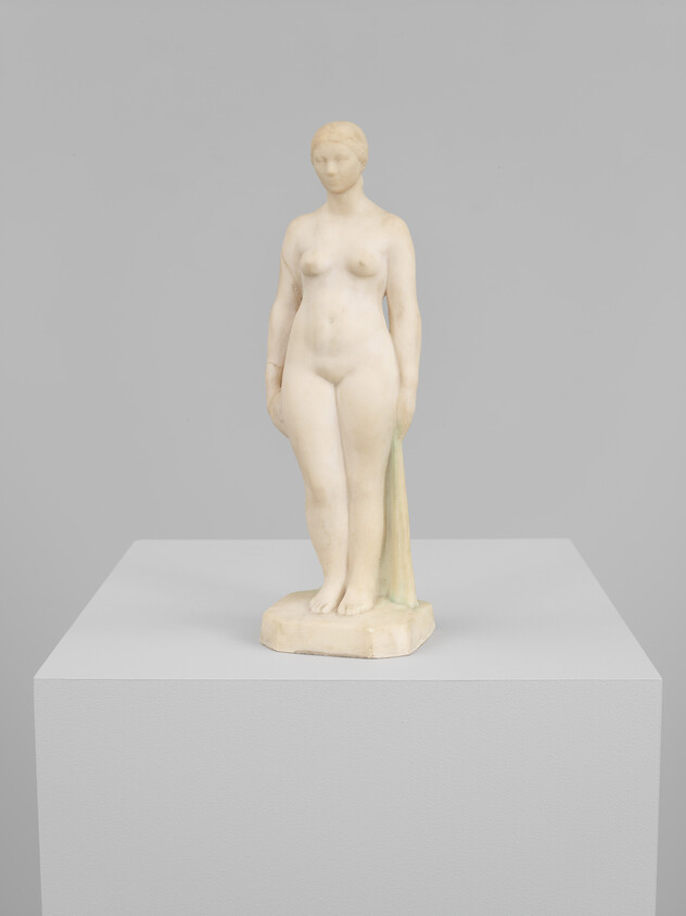 A small classical nude female statue stands on a pedestal holding a draped cloth by her side.