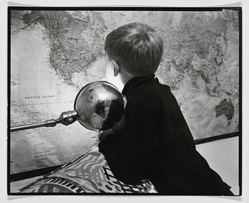 A young boy leans forward looking at a large world map beside a reflective desk lamp.
