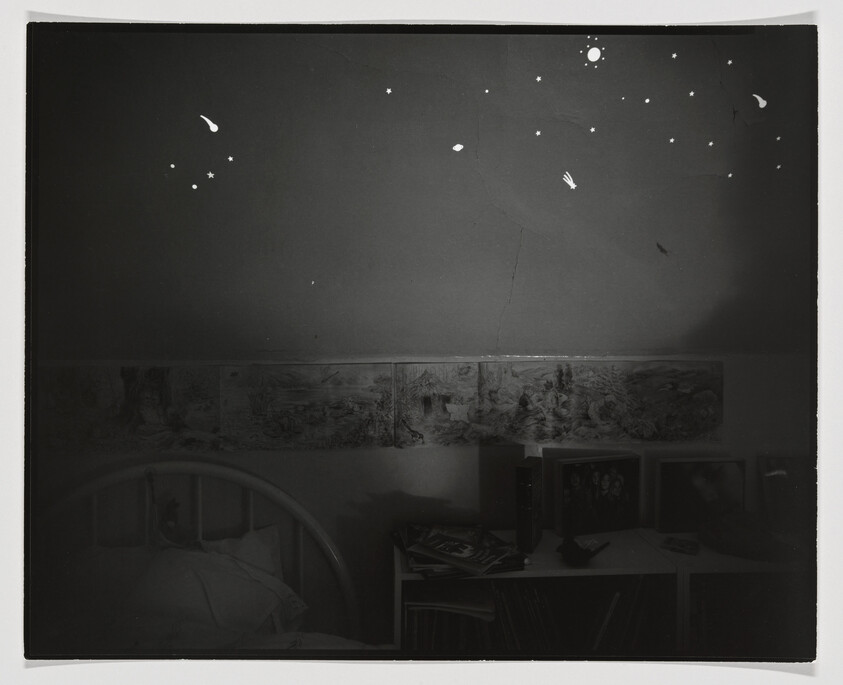 A dark bedroom shows glow-in-the-dark stars on the ceiling above a bed and cluttered shelf.