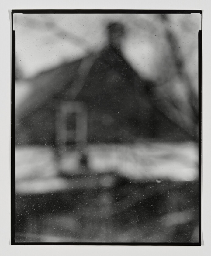 A monochrome, blurry photograph of a house with a prominent gable and a window visible on the front facade. The image has a vintage or aged appearance with dust and scratches on the surface, and the edges of the photo are bordered with black, suggesting it may be an old film photograph. The background is indistinct, possibly showing trees or other vegetation.