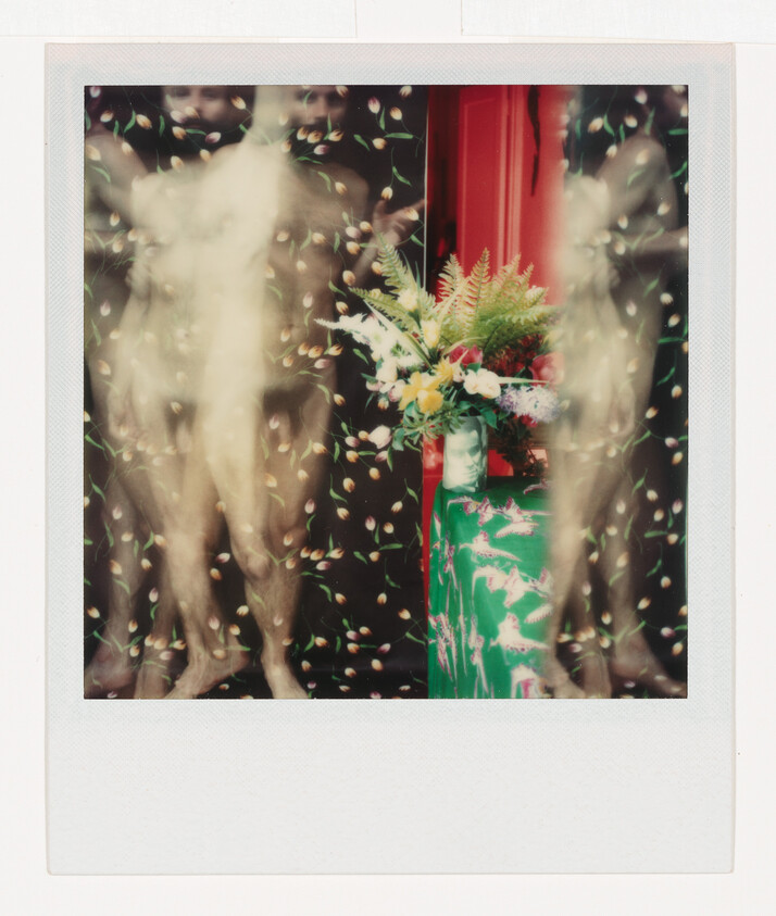 Blurred nude mannequins stand beside a green pedestal holding a colorful bouquet against patterned backdrop.