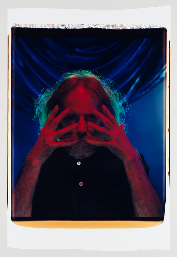 Person with long hair and beard holds hands over their face under dramatic red and blue lighting.