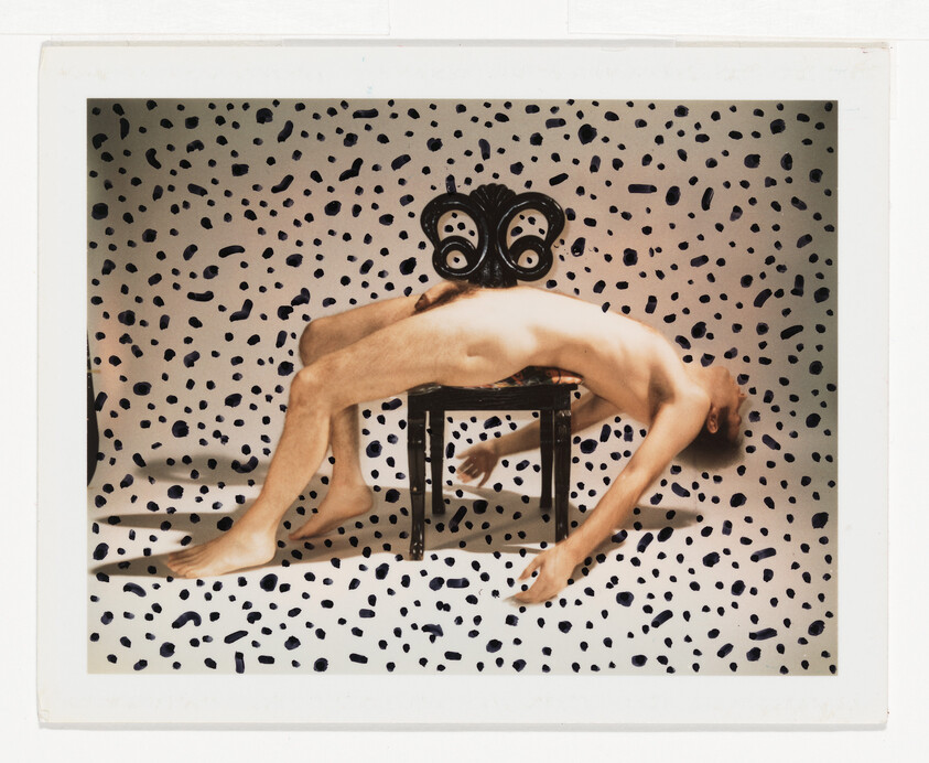A nude person is draped backward over a small ornate chair against a spotted backdrop.