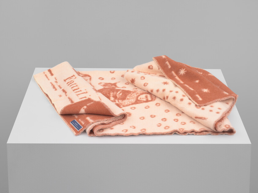 Soft pink and rust patterned wool blanket folded on a white display pedestal.
