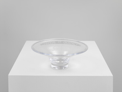 A clear glass shallow bowl sits on a white pedestal with the rim text "CHANGE IT QUICKLY".