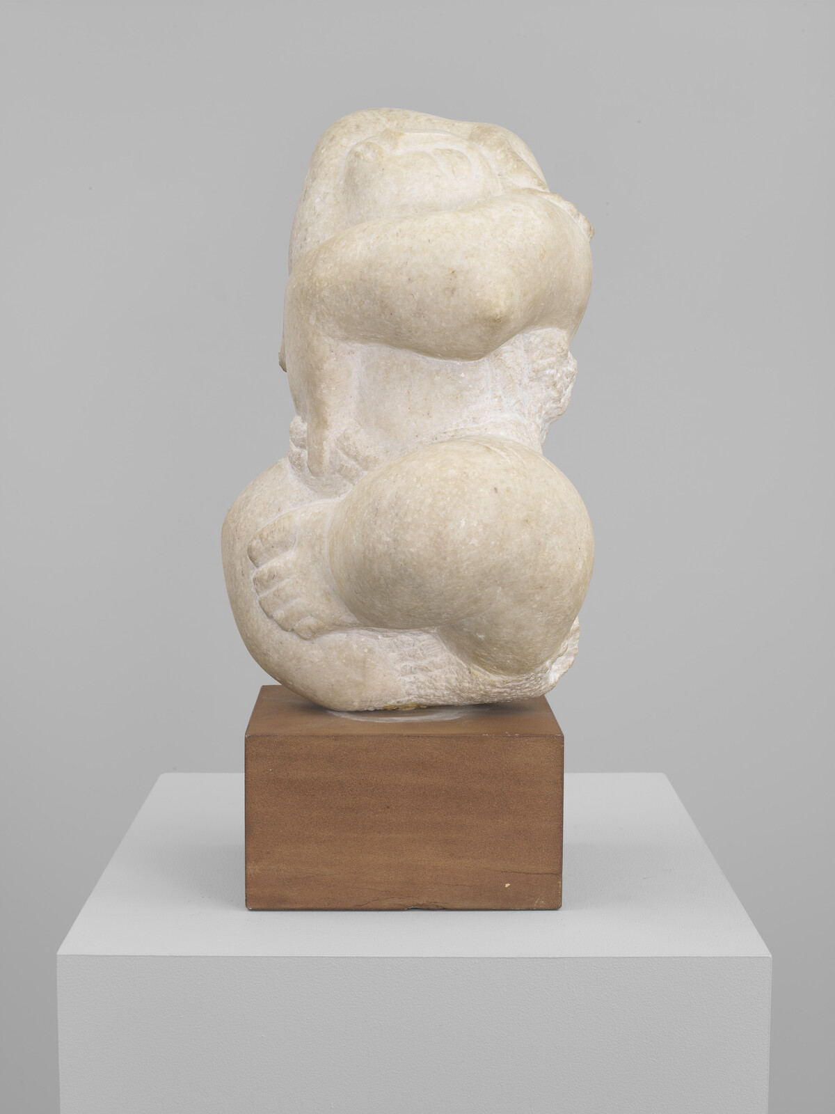 A modern abstract marble sculpture displayed on a wooden pedestal, featuring rounded forms and smooth surfaces, with a neutral background.