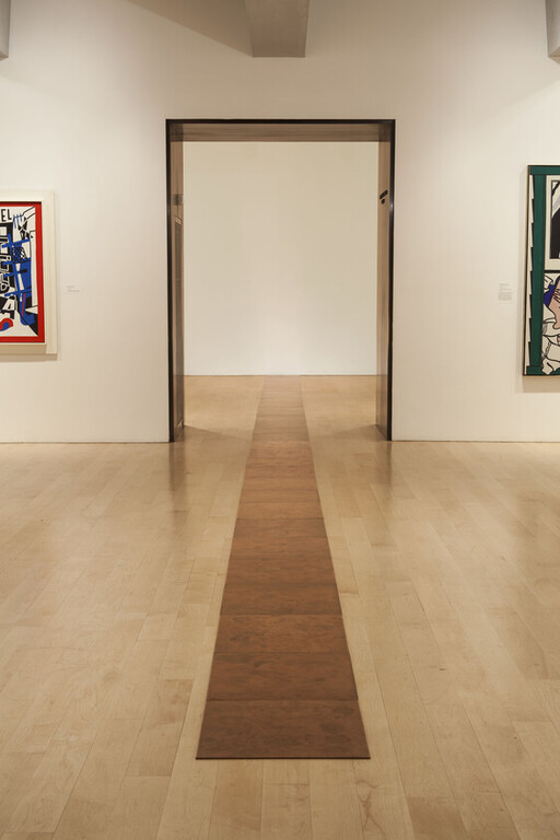 A long narrow wooden strip runs down a gallery floor toward an empty doorway framed by white walls.
