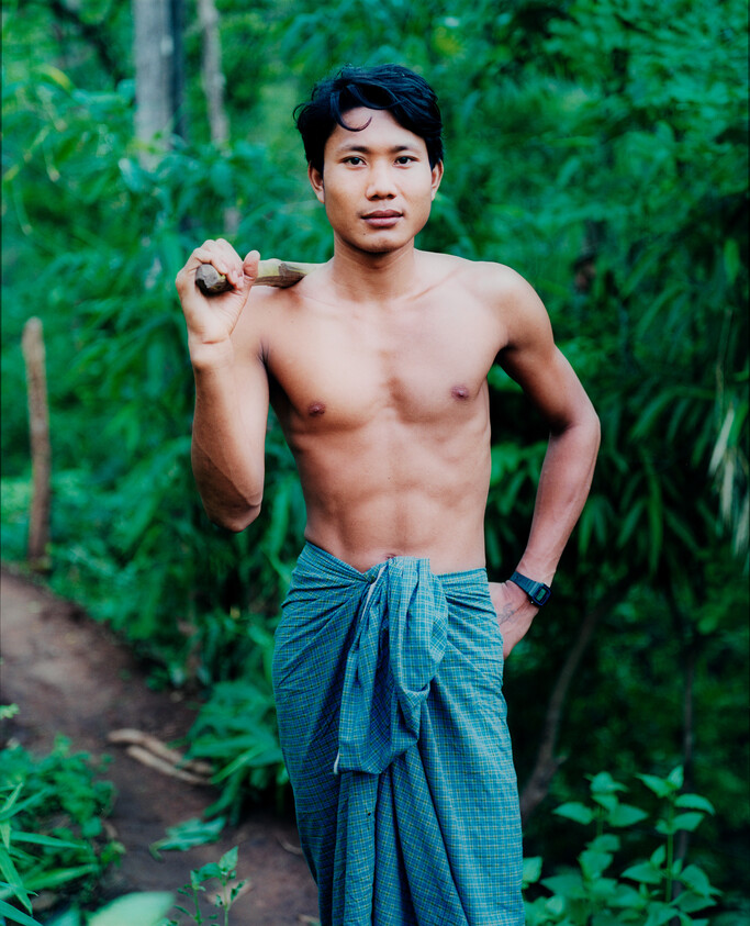 Shirtless young man wearing a sarong carries a stick over his shoulder in a forest.