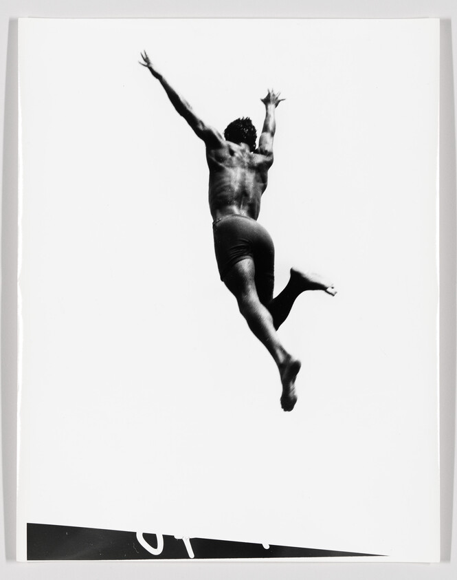 A man in swim trunks leaps upward mid-air with arms outstretched against a white background.