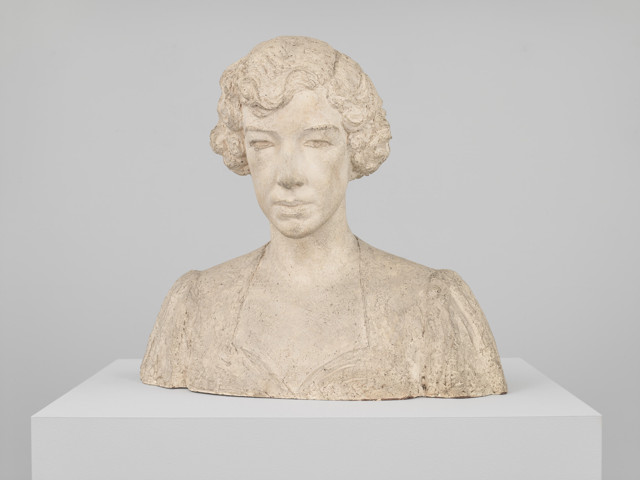 Stone bust of a woman with short curly hair displayed on a white pedestal.