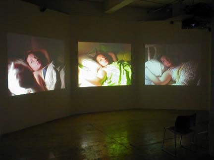 Three projected video screens show a woman sleeping in different poses on a gallery wall.