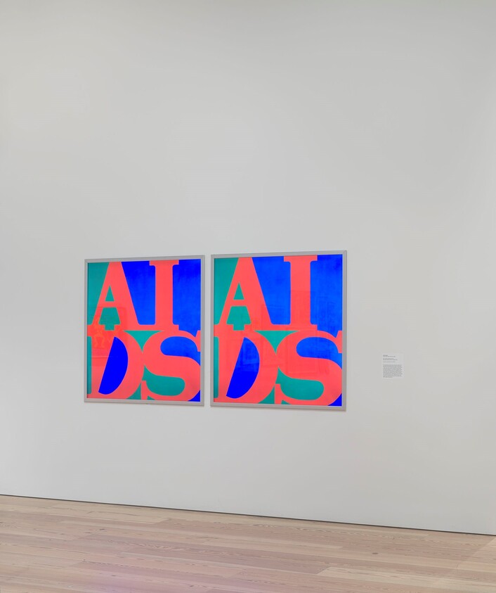 Two framed brightly colored prints with large overlapping letters spelling "AIDS" on a white gallery wall.