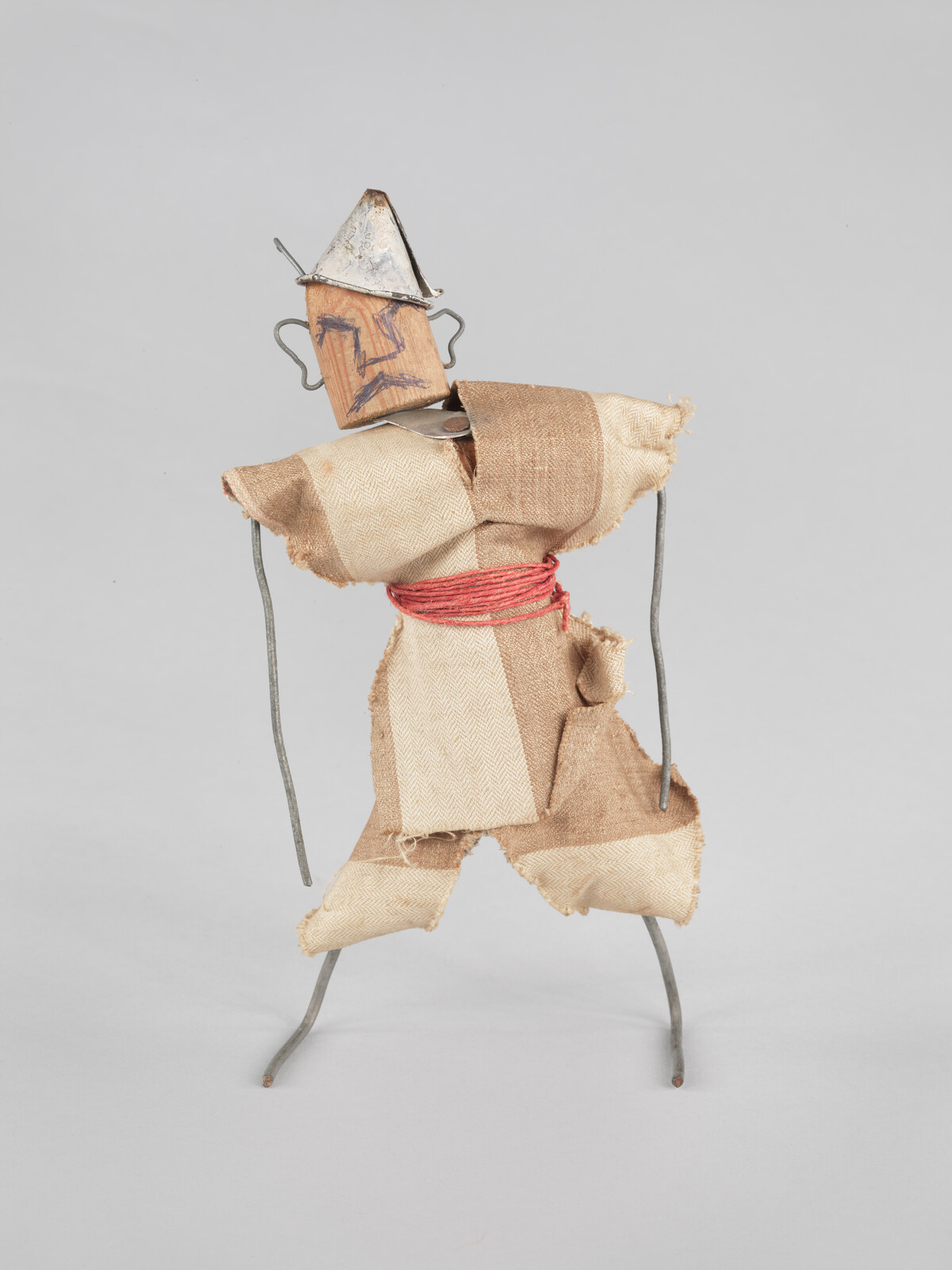 A small figure made of wire, wood, and fabric, wearing a metal hat and red belt.