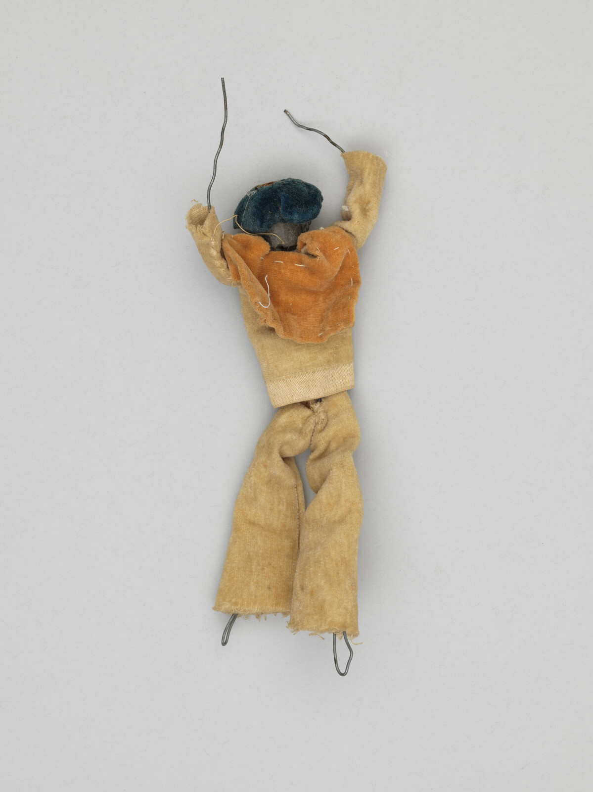 A small, handmade cloth doll with wire arms and legs, wearing a blue hat and orange shirt.