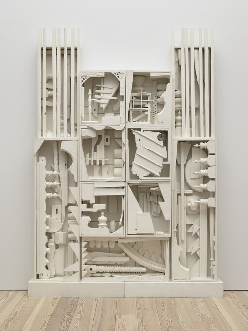 Large white sculpture made from various wooden objects and shapes arranged in a grid against a plain wall.