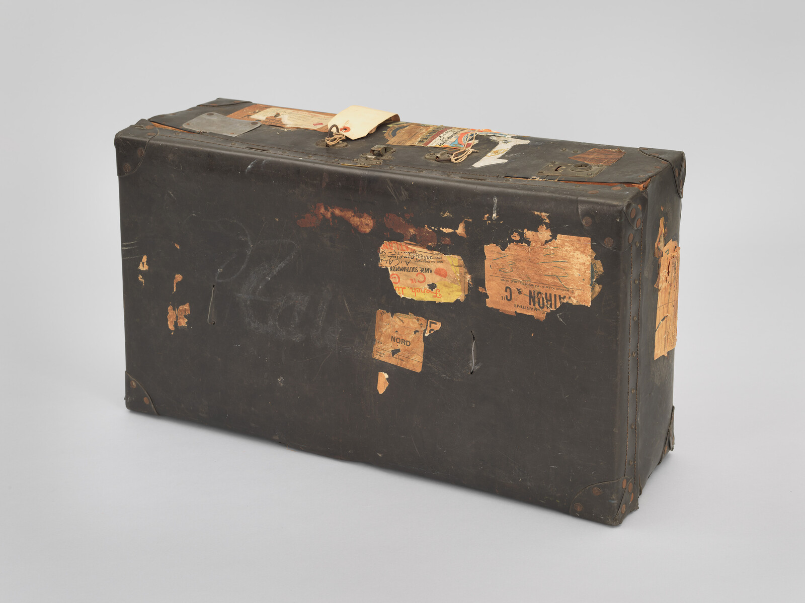 Worn black suitcase with peeling travel stickers and a paper tag attached to the handle.