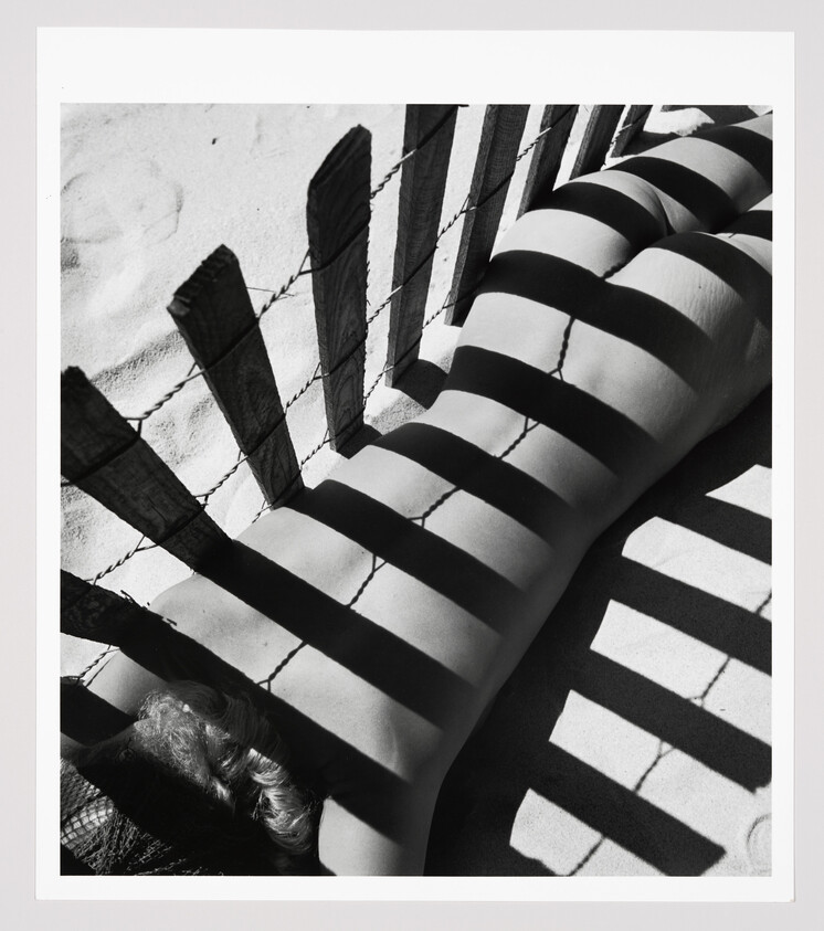 Person lying on sandy beach with a wooden fence casting bold striped shadows across their back.