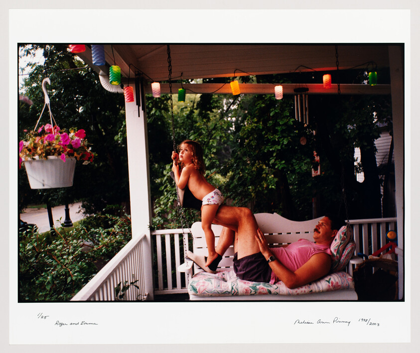 Young child standing on father's legs on a porch swing, reaching up toward hanging lights.