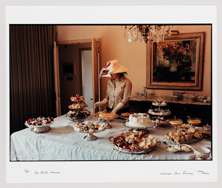 A woman in a decorative hat arranges pastries and cakes on a large tea-party table.