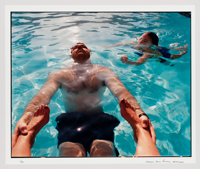 A man floats on his back in a pool holding a woman's feet while a child swims nearby.