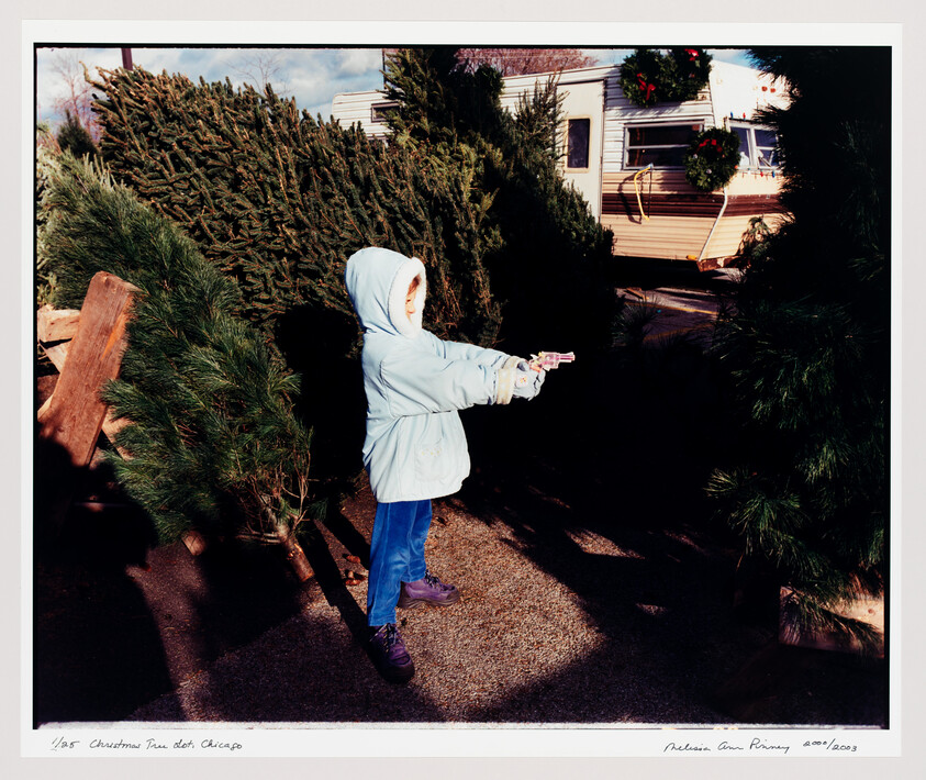 A child in a blue coat points a small pink toy gun among Christmas trees at a lot.