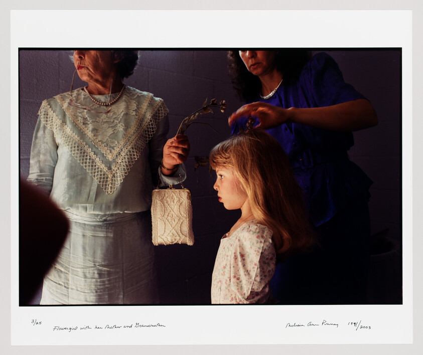 Young girl stands as two women fix her hair and hold a small lace purse.