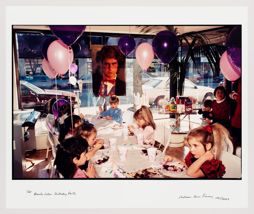 Young children sit around a table eating cake and celebrating a birthday with balloons.