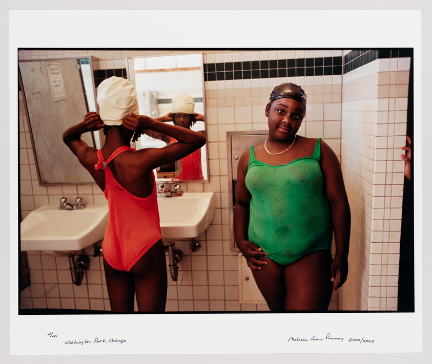 Two girls in a tiled locker room, one adjusting a swim cap while the other stands smiling.