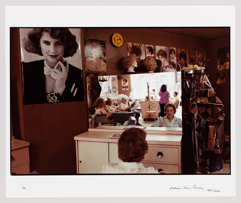 An older woman sits at a salon mirror while other women get their hair dried in the background.