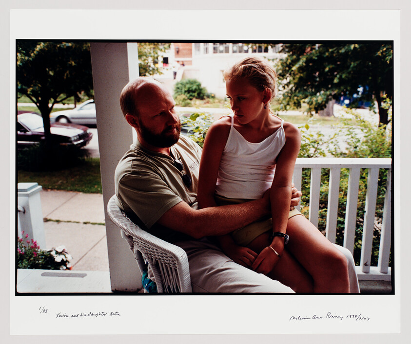 A man sits on a porch holding a young girl on his lap, both looking serious.