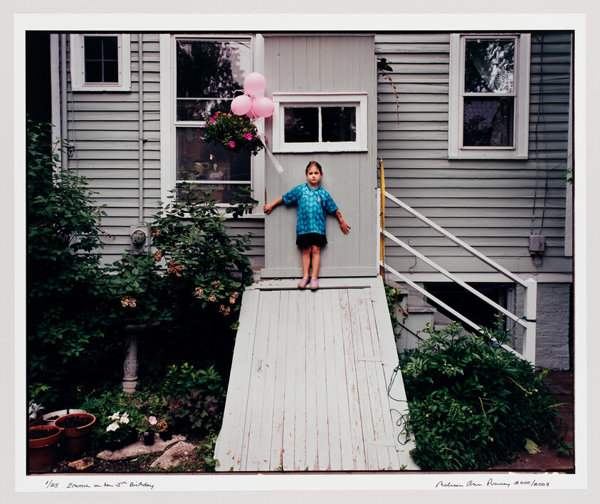 A young girl stands at the top of a wooden ramp outside a gray house holding pink balloons.