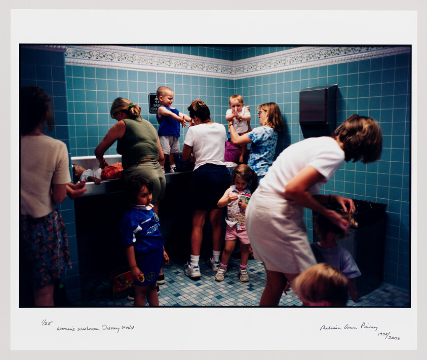 Several women and young children crowded around sinks in a busy tiled restroom, tending to kids.