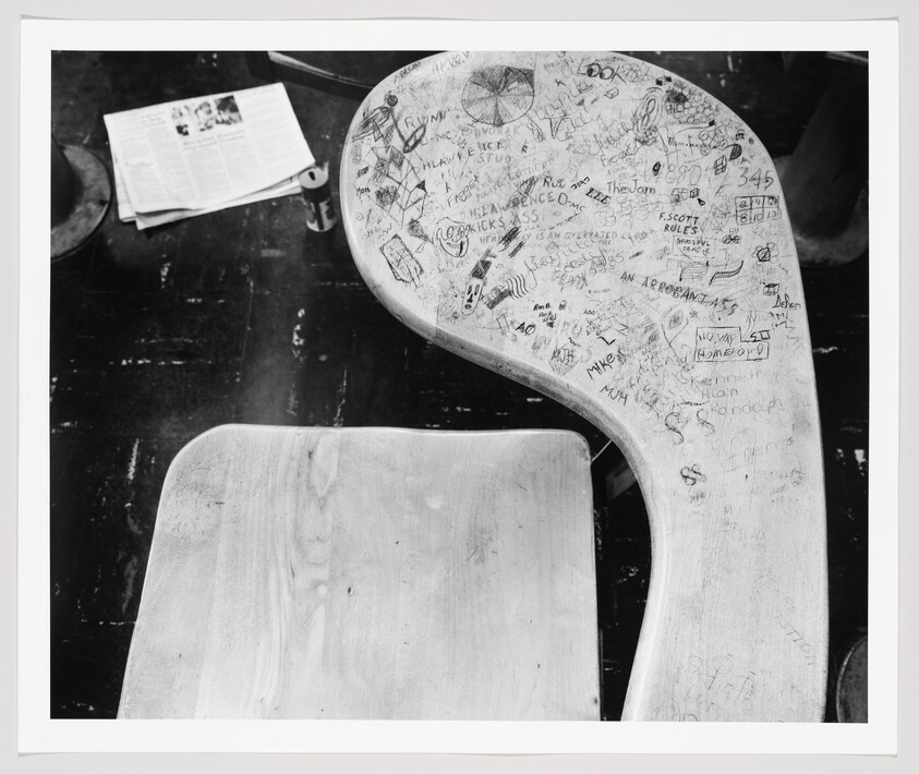 Worn wooden school desk covered in drawings and graffiti, with a newspaper and can on the floor.