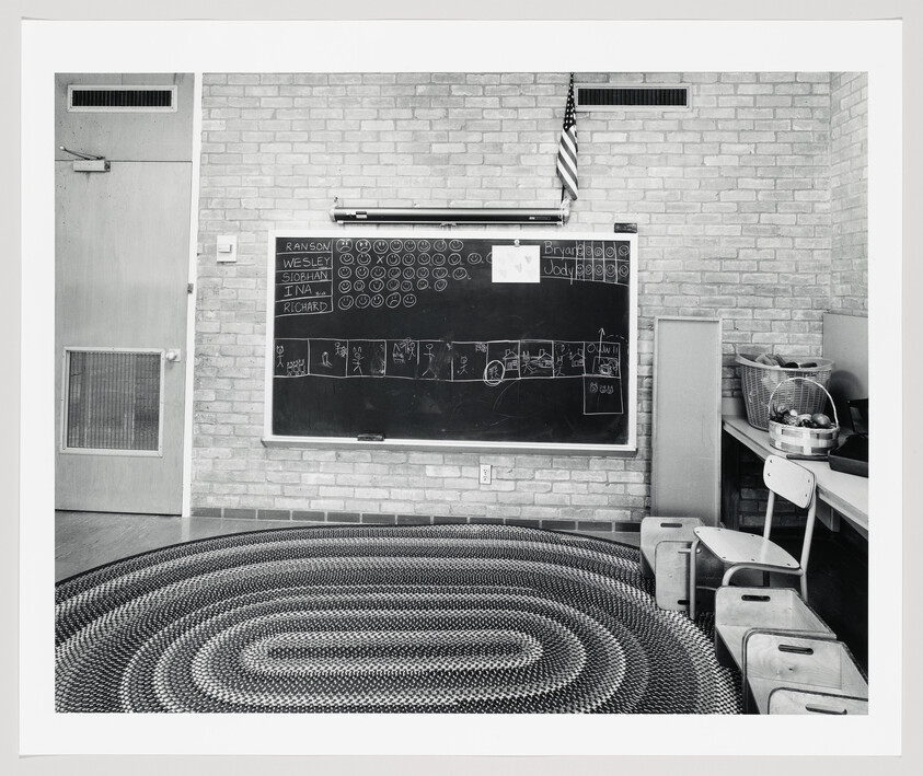 Empty classroom with braided rug, chalkboard showing student names and drawings, chairs and supplies.