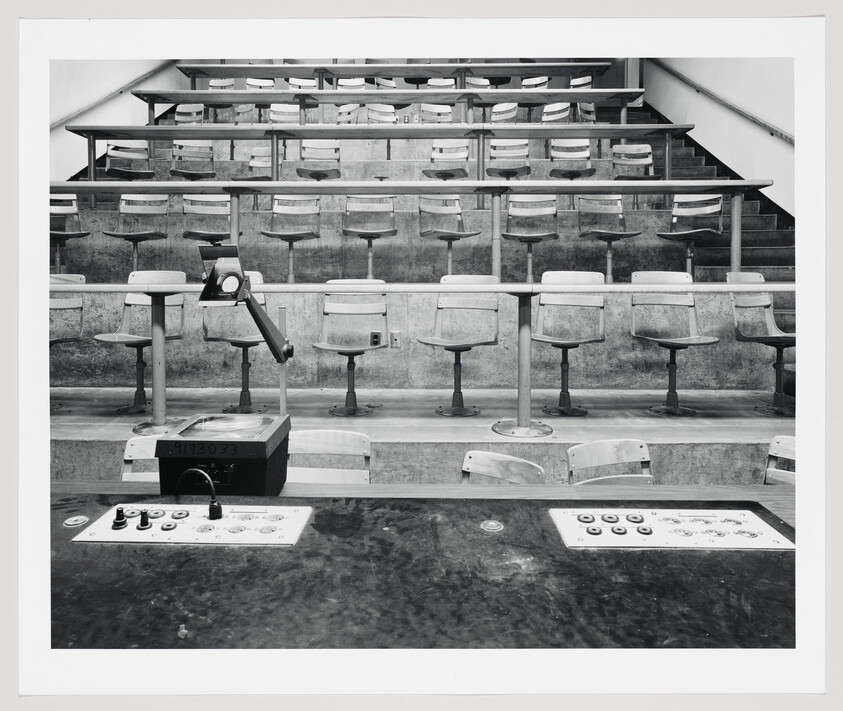 Empty tiered lecture hall with fixed swivel seats, overhead projector, and control panels at front.