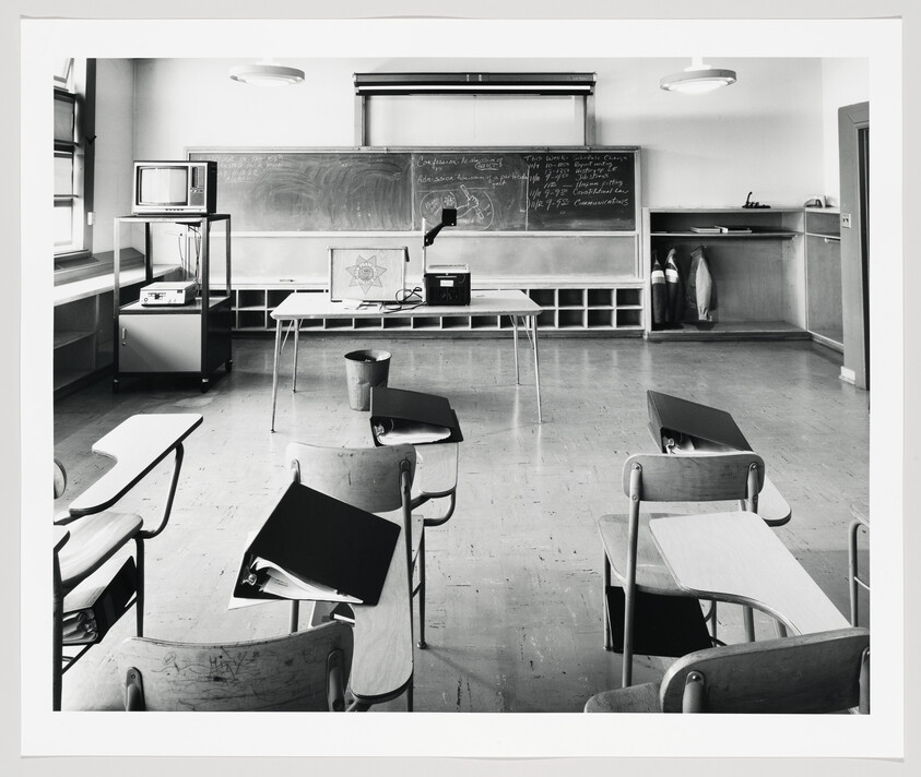 Empty classroom with scattered student desks facing a teacher’s table, overhead projector, and chalkboard.