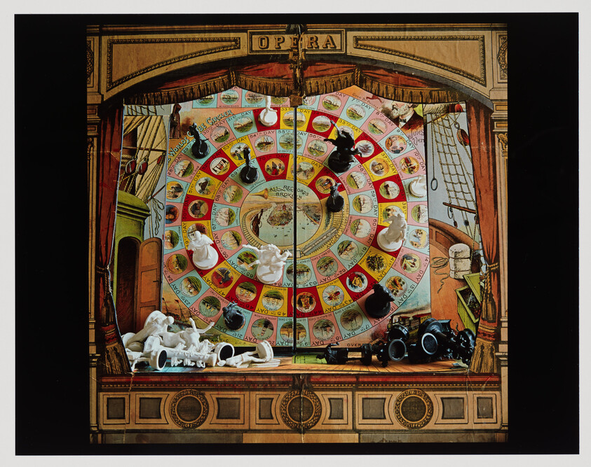 Circular vintage board game set on a small theater stage with scattered white and black playing pieces.